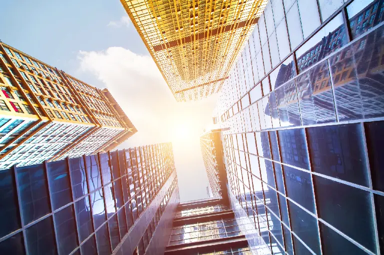 Widok z dołu na budynki biznesowe na tle słonecznego nieba symbolizujące rozbudowę, nowoczesność i wysokie standardy.